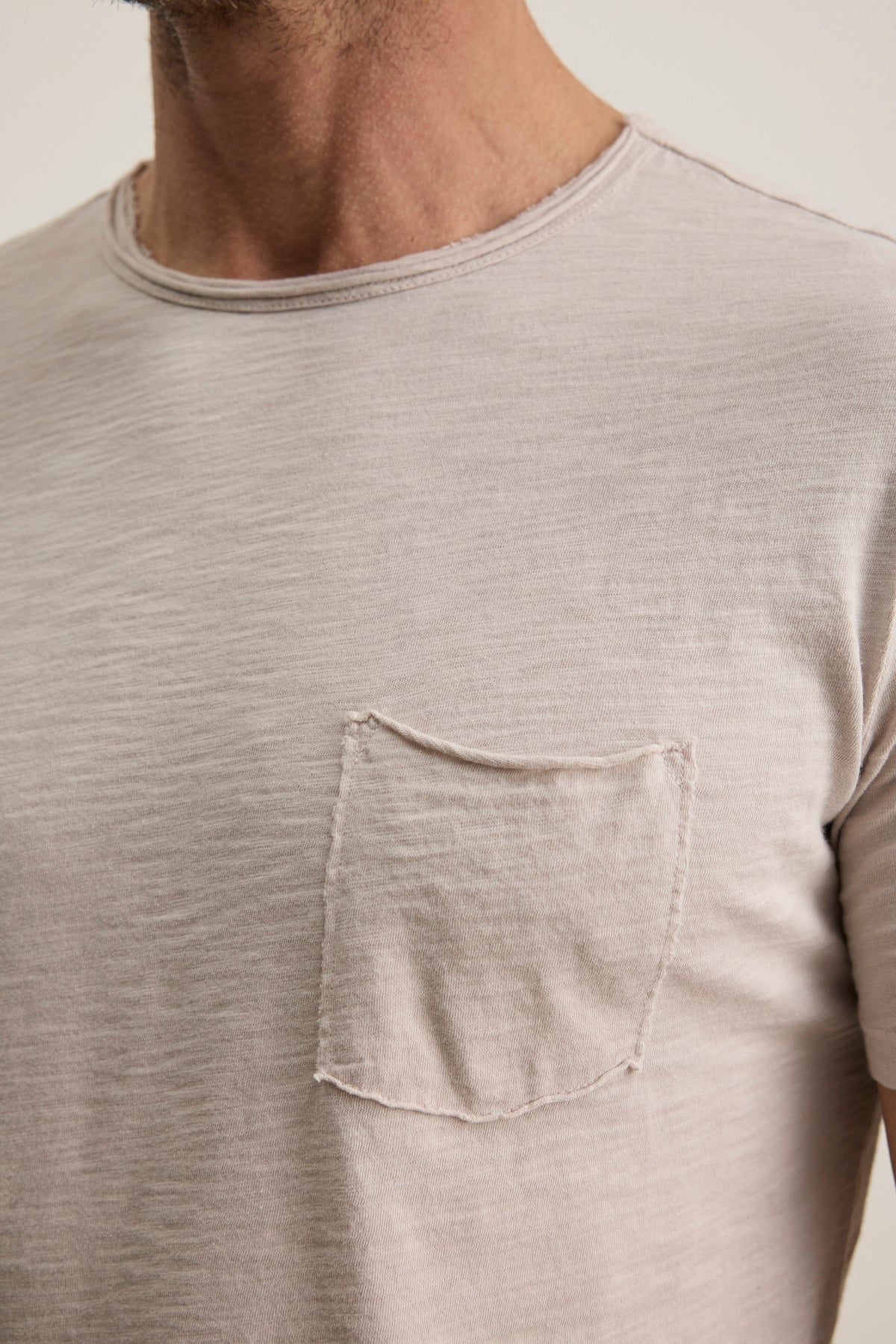   Close-up of a person wearing the Velvet by Graham & Spencer CHAD TEE, a beige T-shirt made from soft Peruvian cotton, featuring a frayed left chest pocket and a classic crew neckline. 