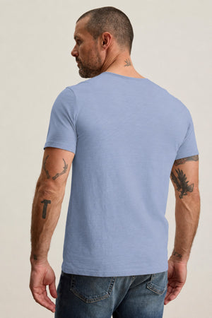 A tattooed man stands with his back to the camera, wearing the CHAD TEE by Velvet by Graham & Spencer—a light blue, ultra-soft Peruvian cotton t-shirt featuring raw edge detailing and a classic crew neckline—paired with blue jeans against a simple, neutral backdrop.