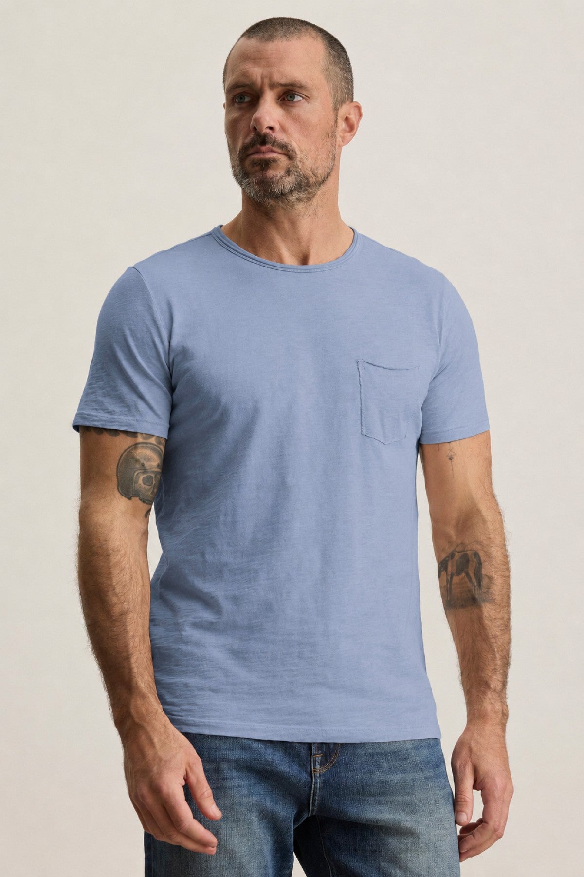 A man with short hair and a beard is wearing the CHAD TEE by Velvet by Graham & Spencer—a light blue, short-sleeved t-shirt made from soft Peruvian cotton with a classic crew neckline—paired with jeans, standing against a plain background. Tattoos are visible on both arms.-41720789500097