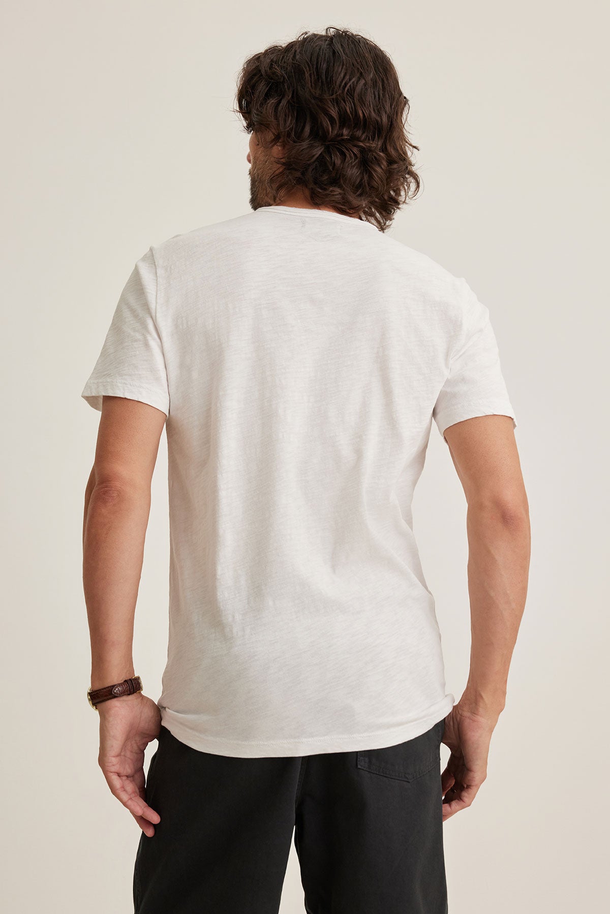   A person stands facing away, hands relaxed at sides, wearing the CHAD TEE by Velvet by Graham & Spencer, a plain white T-shirt made from soft Peruvian cotton, along with dark pants. 