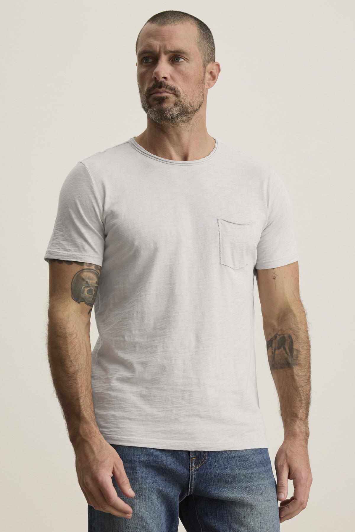   A man with short hair and a beard is wearing the CHAD TEE by Velvet by Graham & Spencer, showcasing his rugged style with tattoos on his arms. 