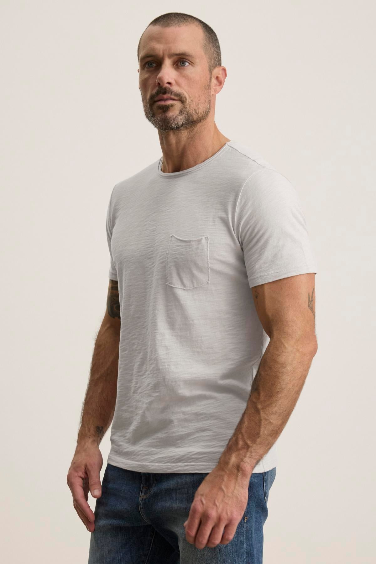   A man with short hair and a beard is wearing the Velvet by Graham & Spencer CHAD TEE in light gray Peruvian cotton, featuring a crew neckline, paired with blue jeans, and standing against a plain background. 