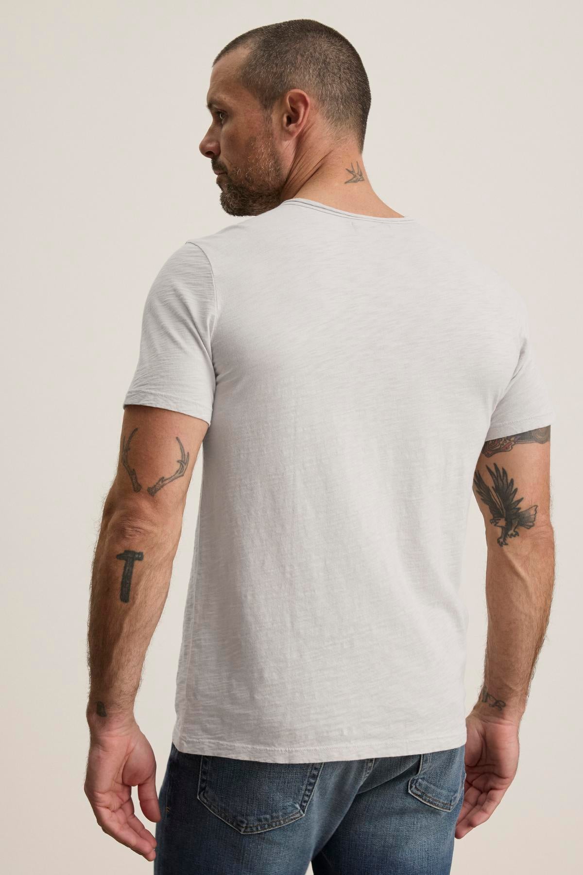   A man with tattoos on his arms stands in a gray Velvet by Graham & Spencer CHAD TEE with a crew neckline and subtle chest pocket, paired with jeans, looking away from the camera. 