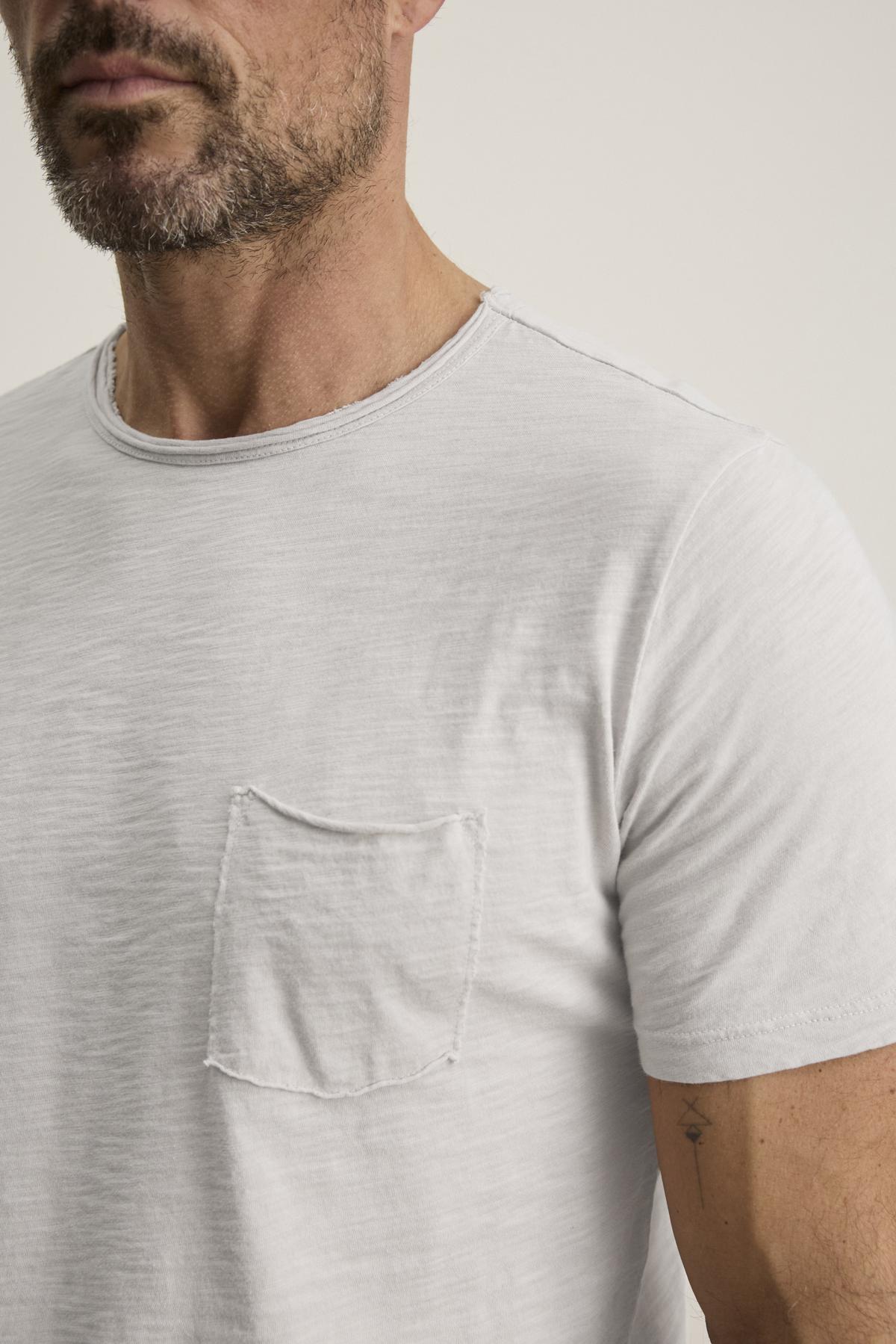   A man is slightly sideways wearing the CHAD TEE by Velvet by Graham & Spencer, a light gray Peruvian cotton T-shirt with a crew neckline and small chest patch pocket. 