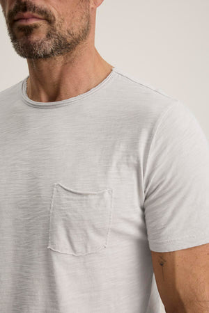 A man is slightly sideways wearing the CHAD TEE by Velvet by Graham & Spencer, a light gray Peruvian cotton T-shirt with a crew neckline and small chest patch pocket.