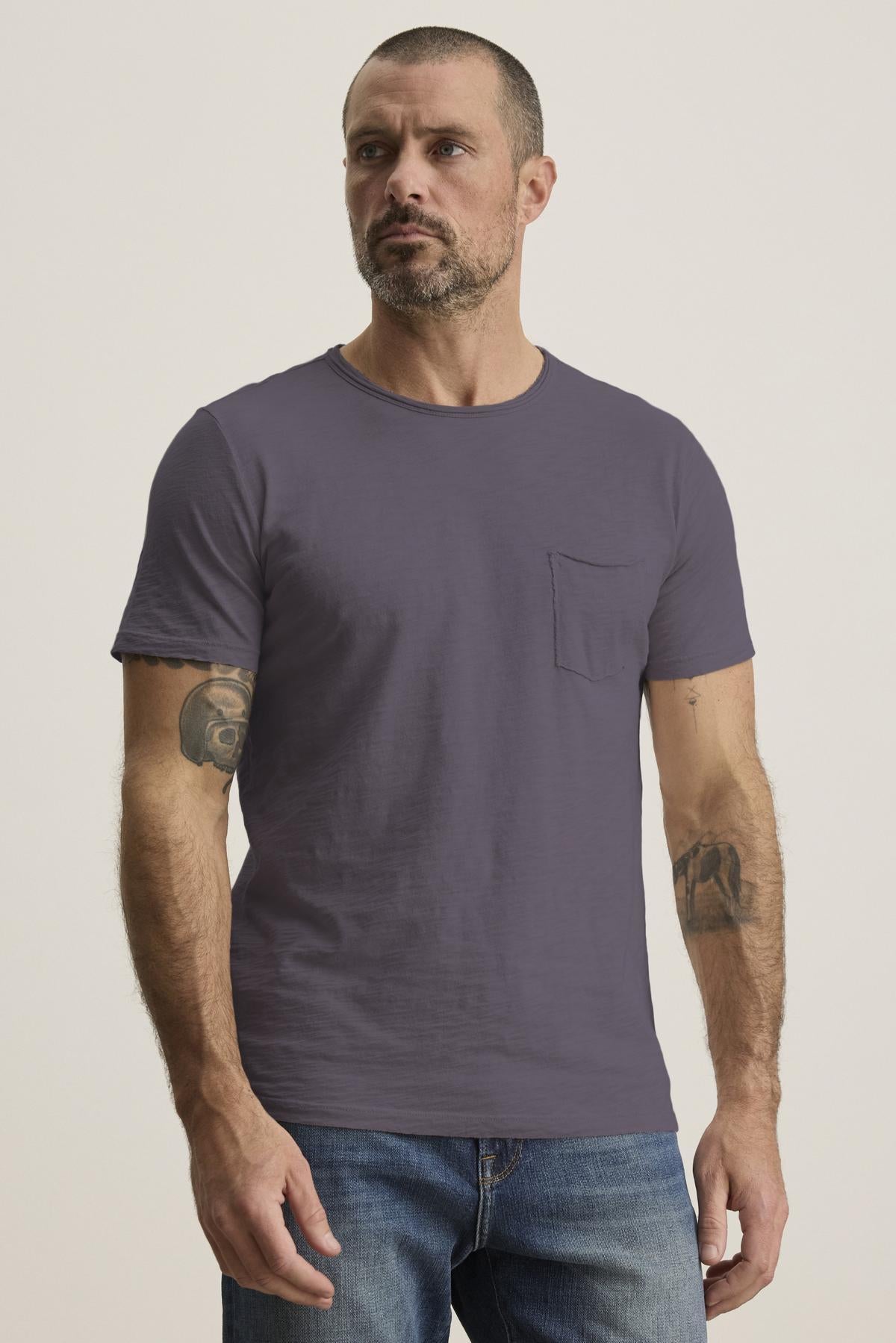   A bearded, tattooed man wears the CHAD TEE, a plain purple crew neck T-shirt from Velvet by Graham & Spencer, made of soft Peruvian cotton, paired with jeans. 