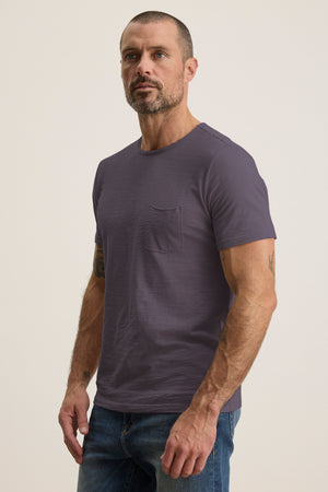 A person with short hair and a beard wears Velvet by Graham & Spencer's CHAD TEE in gray, made from soft Peruvian cotton, with a crew neckline and chest patch pocket, paired with jeans against a neutral background.