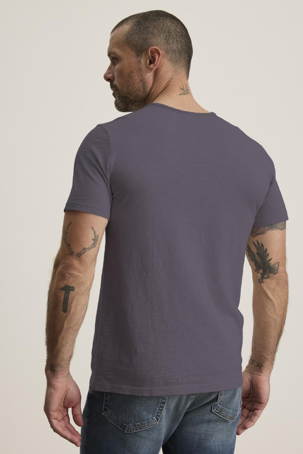   A man with short hair and tattoos on his arms and neck wears the CHAD TEE in gray, featuring a crew neckline, by Velvet by Graham & Spencer paired with jeans, viewed from behind. 