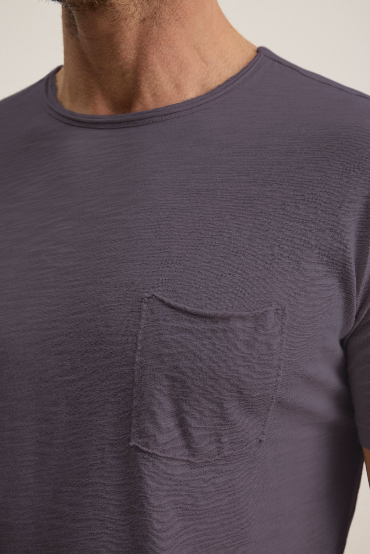   A close-up shows a person wearing the Velvet by Graham & Spencer's CHAD TEE, a plain dark gray T-shirt made of soft Peruvian cotton, featuring a classic crew neckline and a small chest pocket. The image captures the upper torso and part of the neck. 