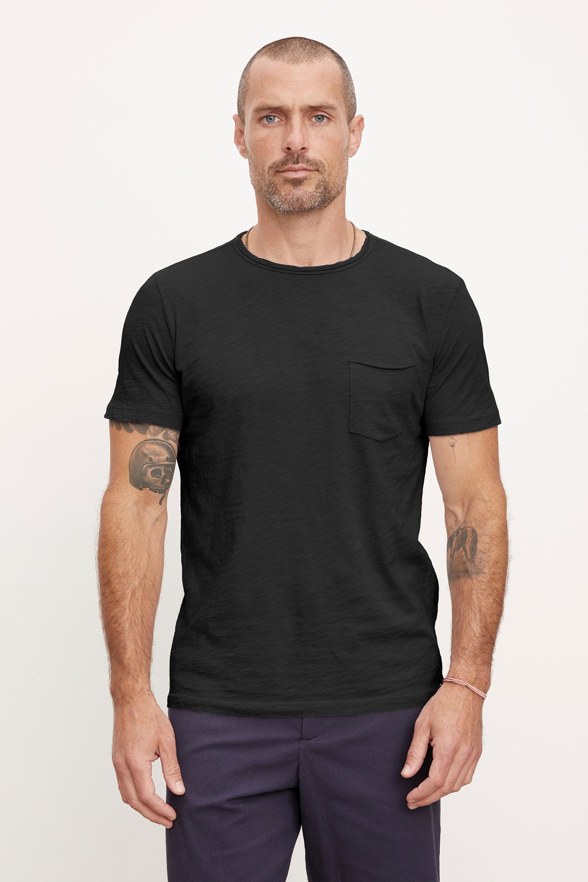   Man in black CHAD TEE with raw-edge details by Velvet by Graham & Spencer and dark trousers, standing straight and looking at the camera, with tattooed arms. 
