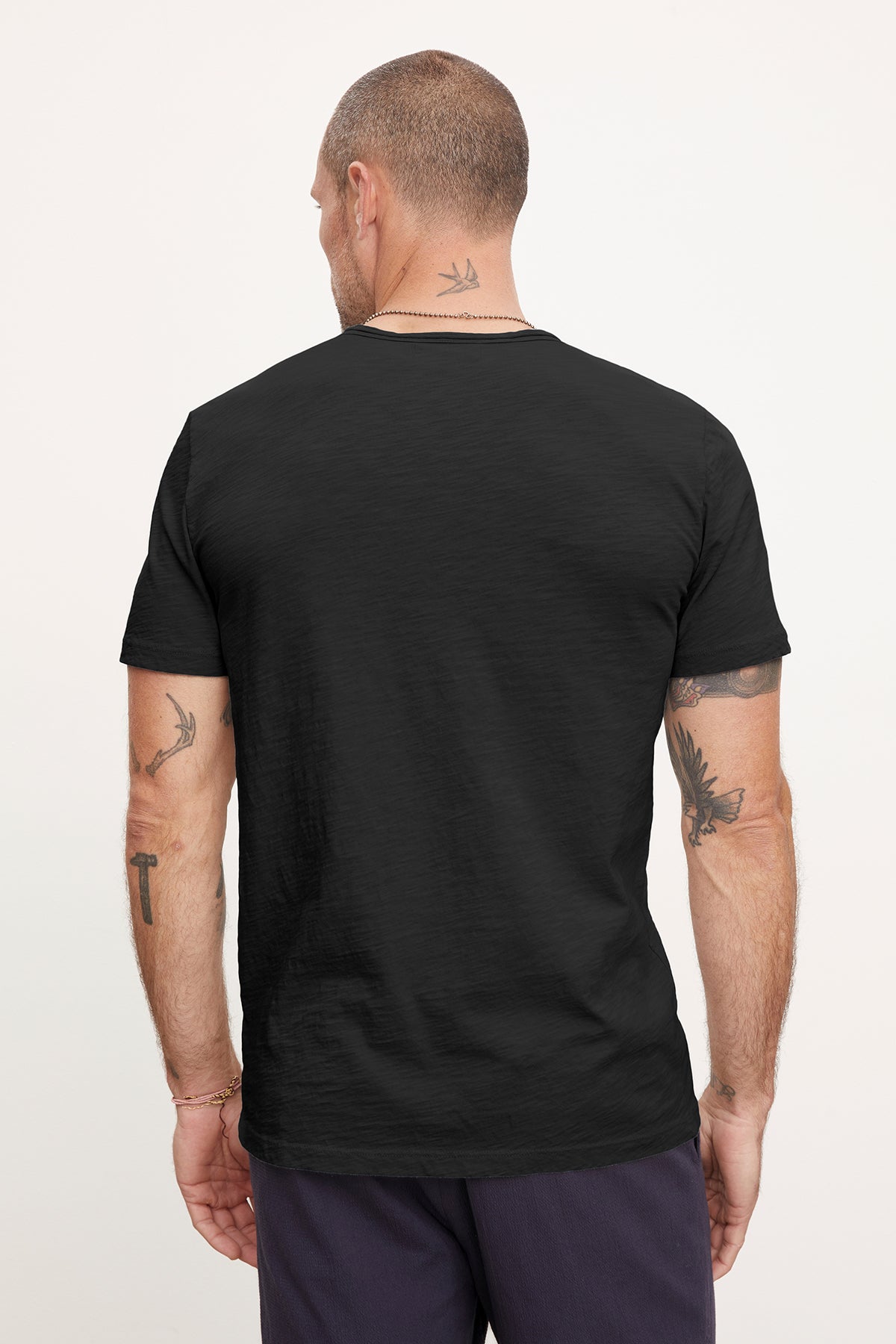 A man seen from the back wearing a Velvet by Graham & Spencer CHAD TEE with raw-edge details and purple pants, displaying various tattoos on his arms and neck.-36909374832833