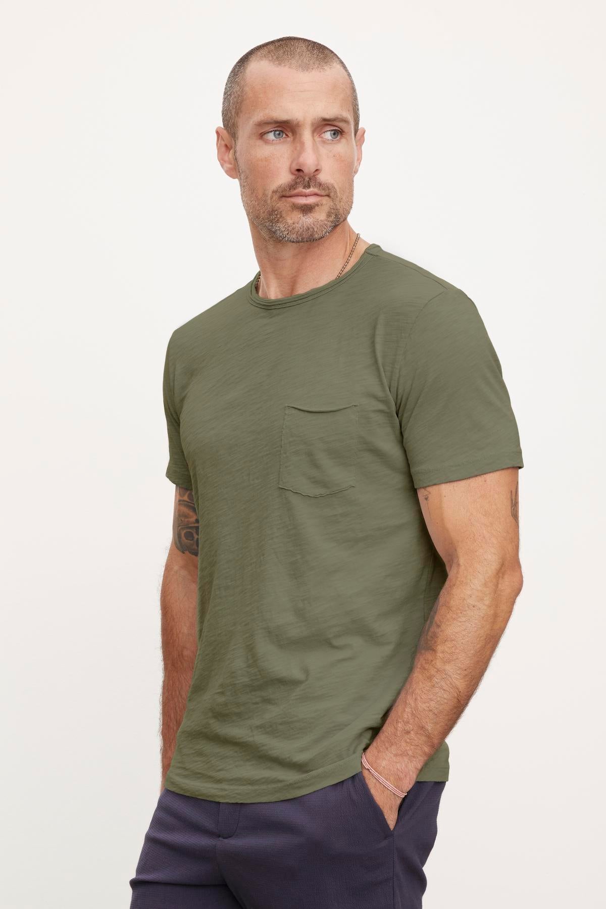   A man with a cropped haircut and beard, wearing the CHAD TEE by Velvet by Graham & Spencer with a classic crew neckline and dark pants, stands looking off to the side. His left arm has visible tattoos, while the textured cotton slub of his shirt adds a unique touch. 