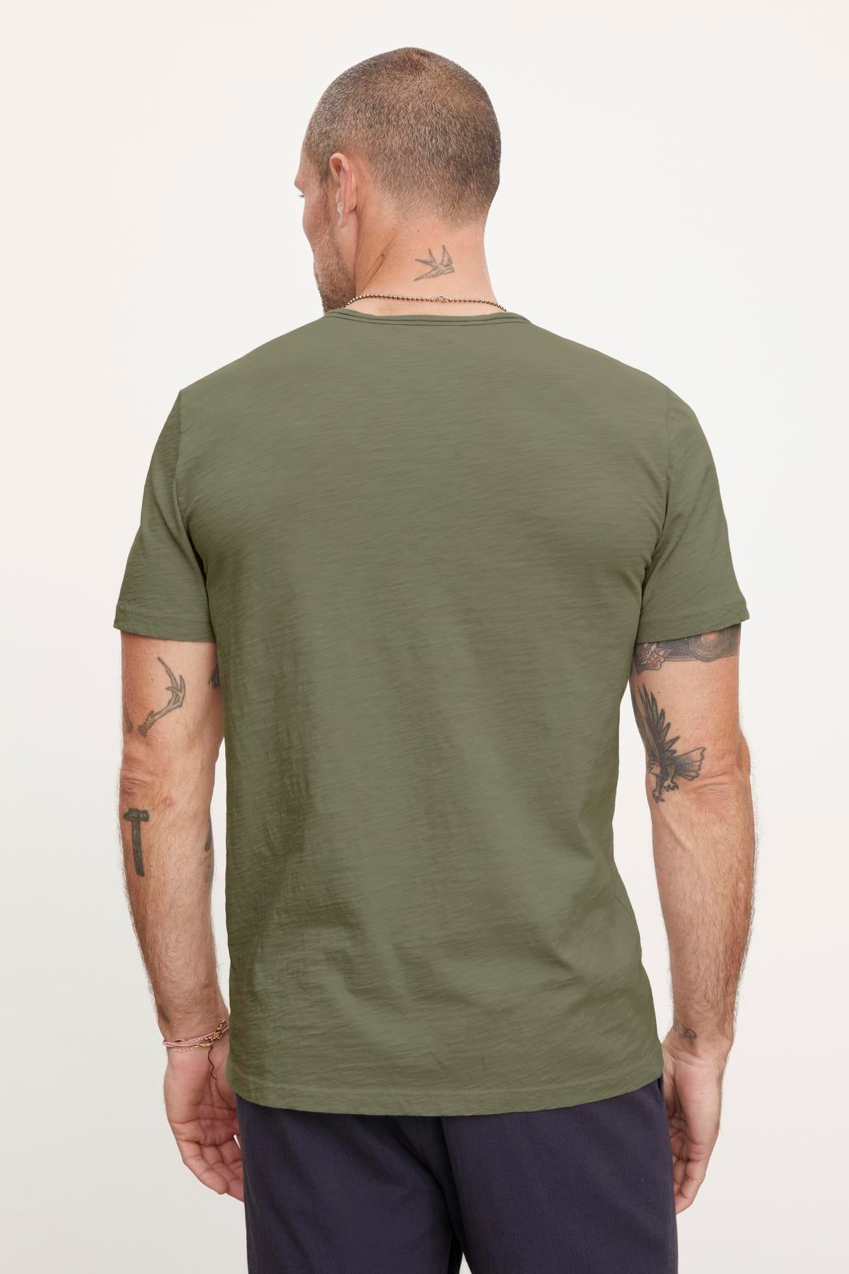   Man with a shaved head and tattoos on his neck and arms, sporting an olive green short-sleeve CHAD TEE made from textured cotton slub by Velvet by Graham & Spencer, and dark pants, shown from the back. 