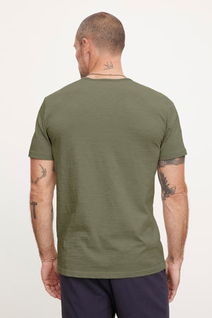 Man with a shaved head and tattoos on his neck and arms, sporting an olive green short-sleeve CHAD TEE made from textured cotton slub by Velvet by Graham & Spencer, and dark pants, shown from the back.