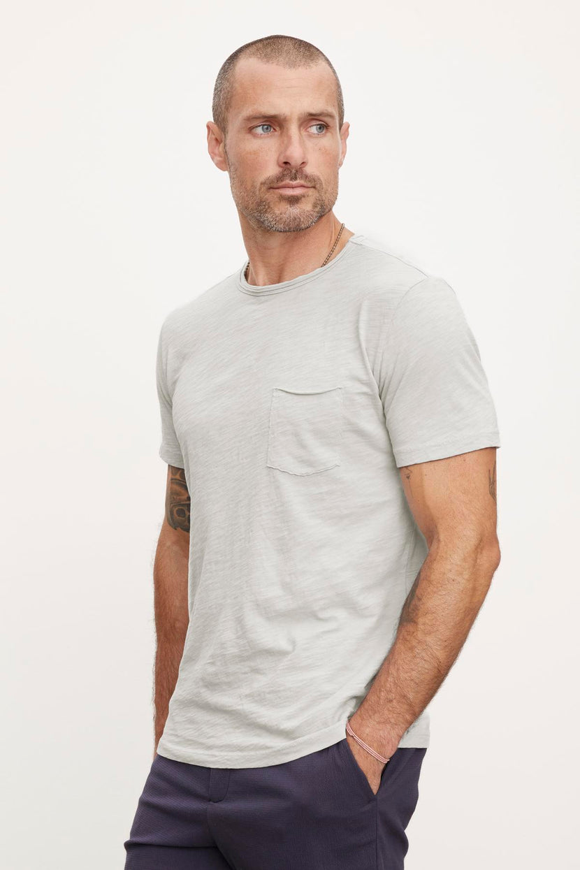 A man with a beard and short hair is wearing a light gray textured cotton **CHAD TEE** by **Velvet by Graham & Spencer** with raw edge details and dark pants, standing against a plain background.