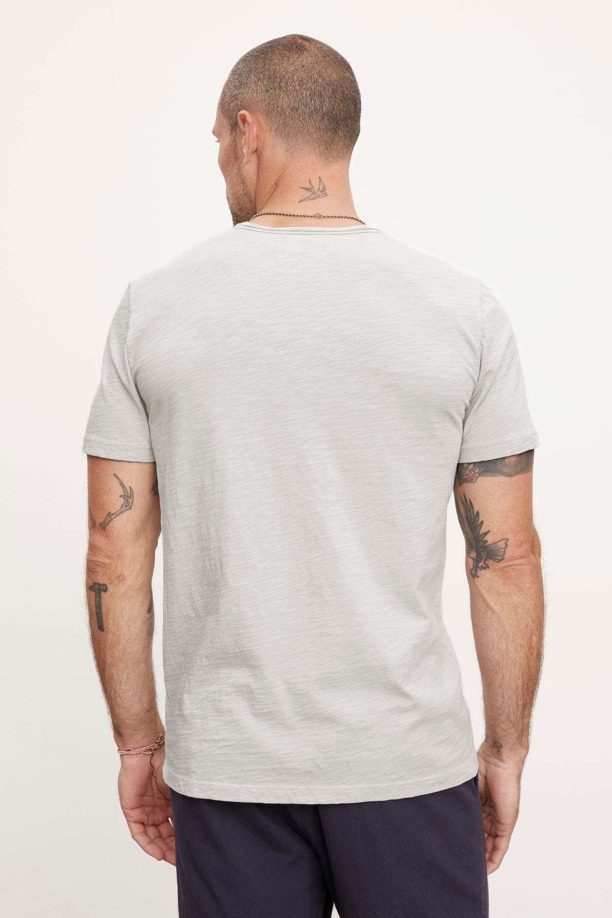 Man seen from the back, standing and wearing a Velvet by Graham & Spencer CHAD TEE with raw edge details and dark pants. His arms and neck have tattoos.-37386059448513