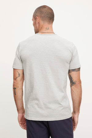 Man seen from the back, standing and wearing a Velvet by Graham & Spencer CHAD TEE with raw edge details and dark pants. His arms and neck have tattoos.