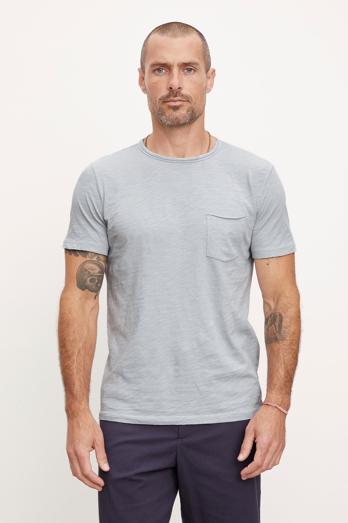 A man with a neutral expression wearing a Velvet by Graham & Spencer CHAD TEE with raw-edge details and dark pants, standing against a plain white background. He has visible tattoos on his arms.-36890799014081