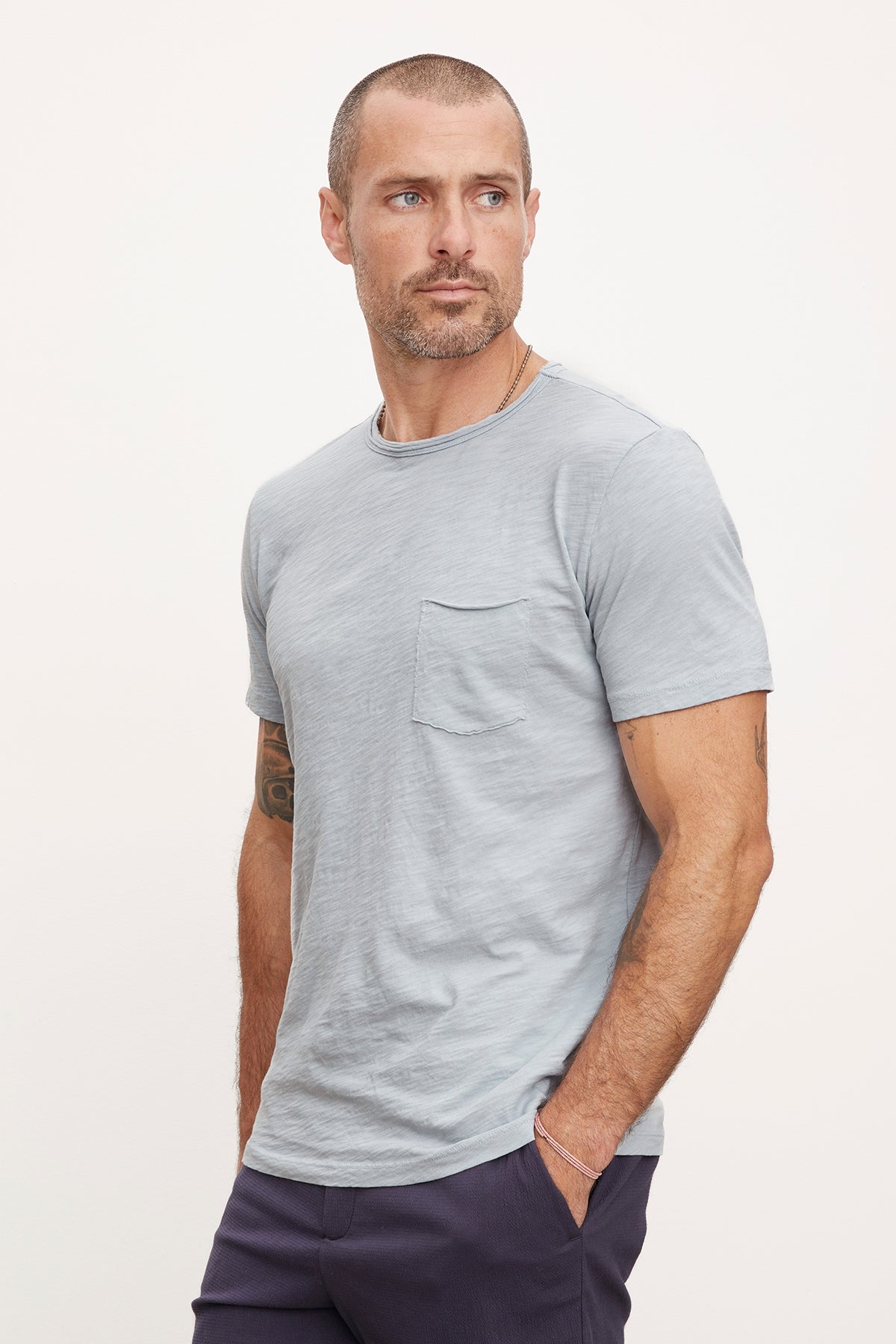 A man in a gray textured cotton slub CHAD TEE by Velvet by Graham & Spencer and dark pants standing against a white background, looking to the side with a serious expression.-36890799046849