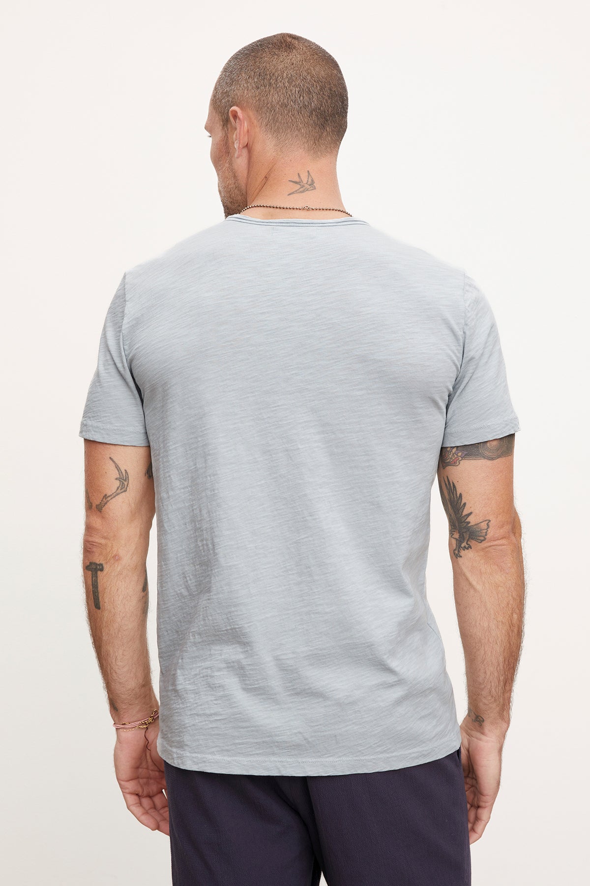Man standing with his back to the camera showcasing a Velvet by Graham & Spencer CHAD TEE and navy shorts, displaying visible tattoos on his arms and neck.-36890799079617