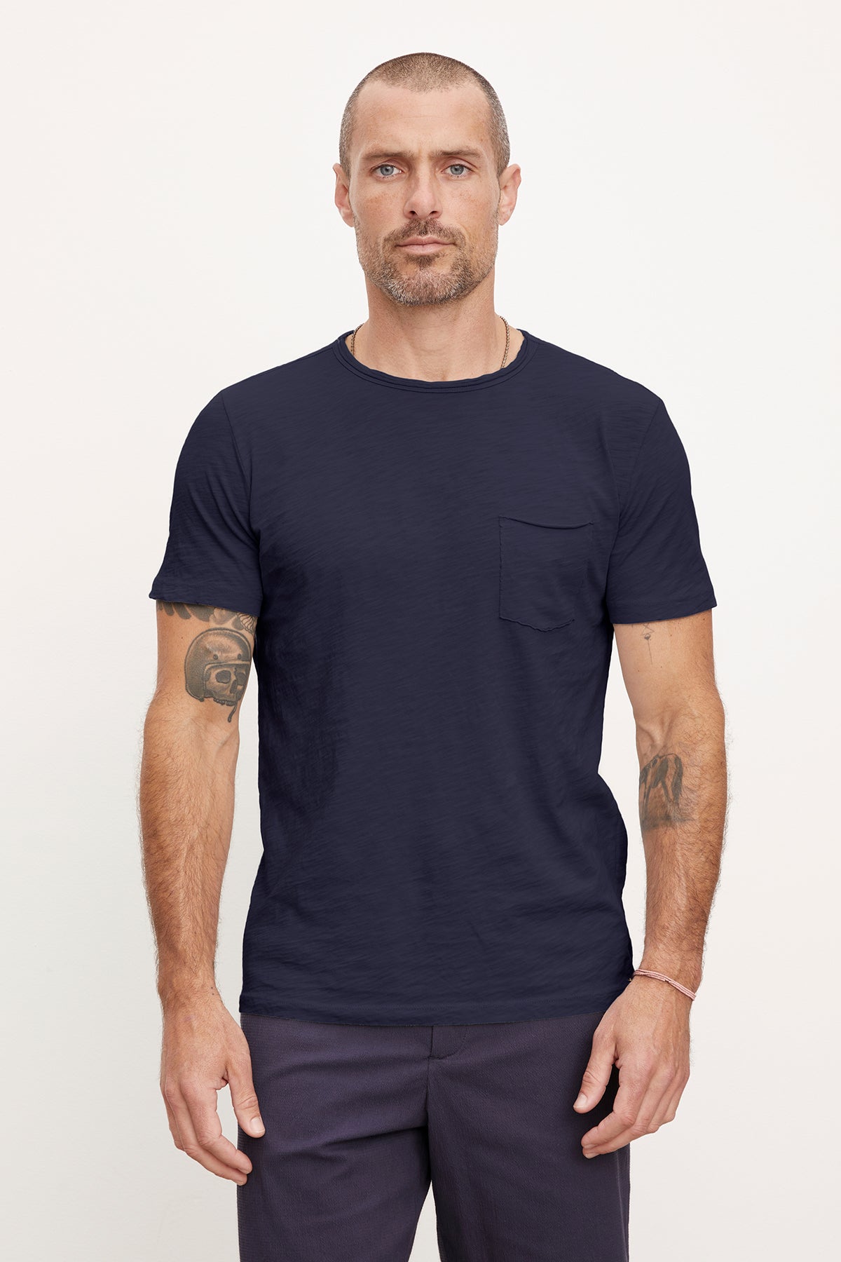   A man with short hair and tattoos on his arms, wearing a dark blue textured cotton slub CHAD TEE by Velvet by Graham & Spencer and navy blue pants, standing against a plain white background. 