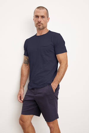 A man in a navy blue CHAD TEE by Velvet by Graham & Spencer with raw-edge details and matching shorts stands against a plain white background, looking directly at the camera.