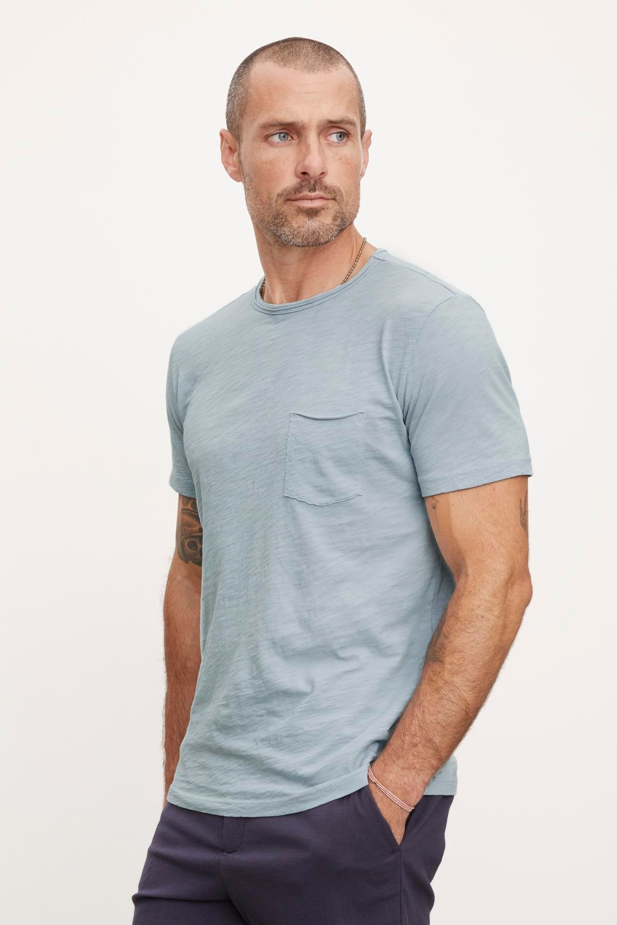   A person with short hair and a beard wears a light blue CHAD TEE by Velvet by Graham & Spencer with raw edge details and dark pants, looking to the side. The classic crew neckline t-shirt has a chest pocket made of textured cotton slub. The background is plain and light-colored. 