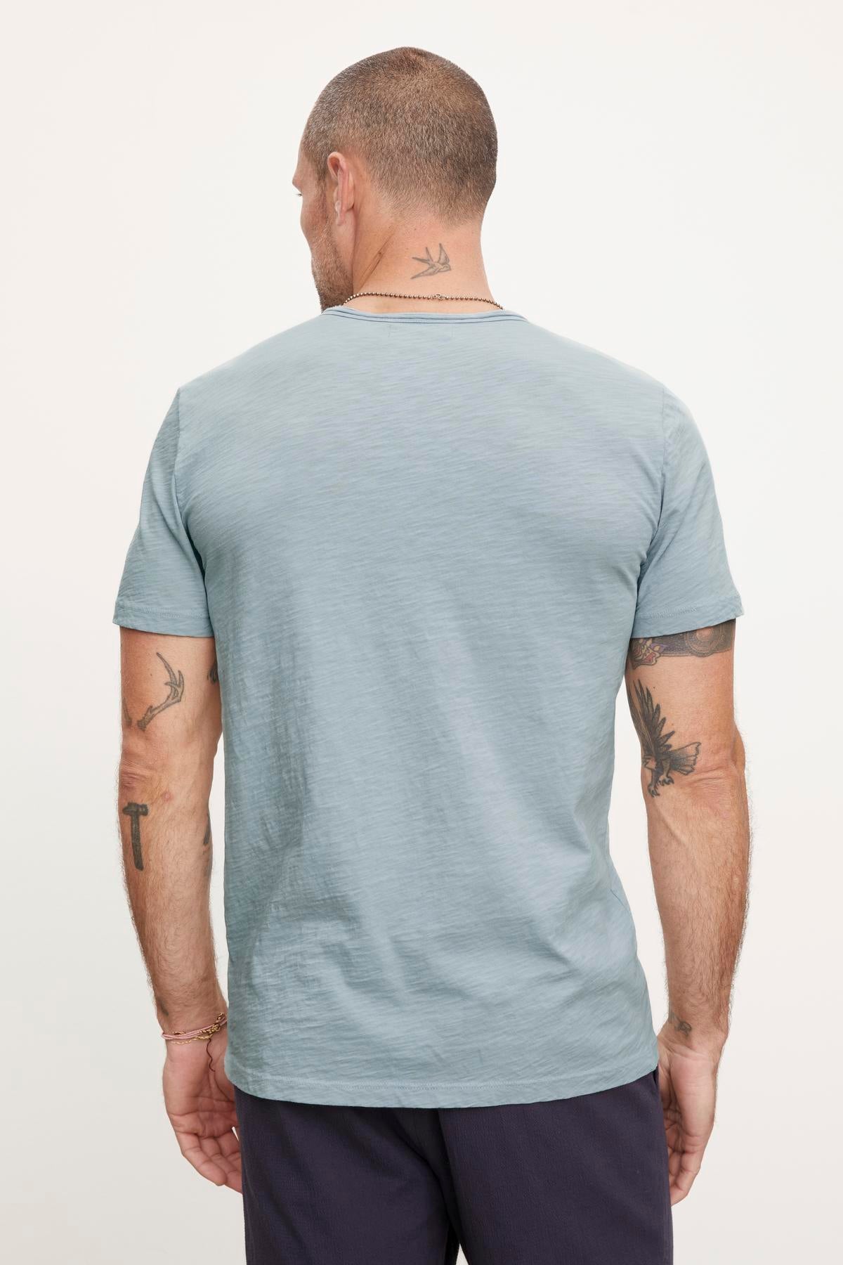   A man with short hair and tattoos on both arms is shown from the back, wearing a light blue CHAD TEE by Velvet by Graham & Spencer made of textured cotton slub with raw edge details, paired with dark pants. 