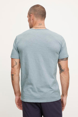 A man with short hair and tattoos on both arms is shown from the back, wearing a light blue CHAD TEE by Velvet by Graham & Spencer made of textured cotton slub with raw edge details, paired with dark pants.