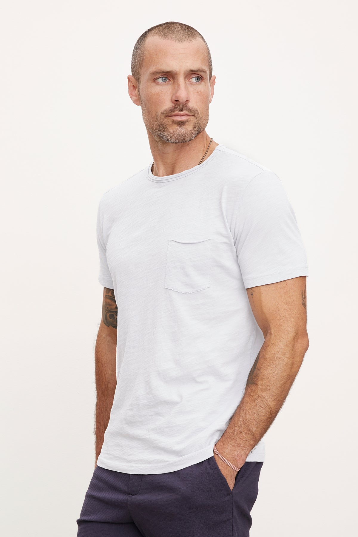   A man with a beard and tattoos, wearing a white textured cotton slub CHAD TEE by Velvet by Graham & Spencer and navy trousers, stands with a serious expression looking to his left. 