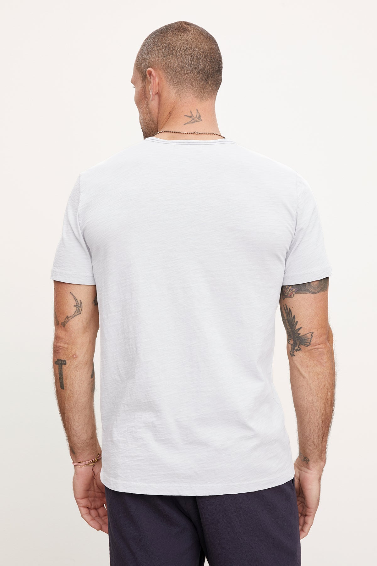   A man viewed from behind, wearing a white CHAD TEE with raw-edge details by Velvet by Graham & Spencer and navy pants, showcasing tattoos on his arms and a neck tattoo. 