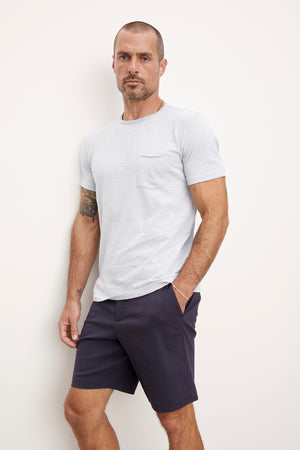 A man in a white textured cotton slub CHAD TEE from Velvet by Graham & Spencer and navy shorts standing with his hands on his hips against a plain background.