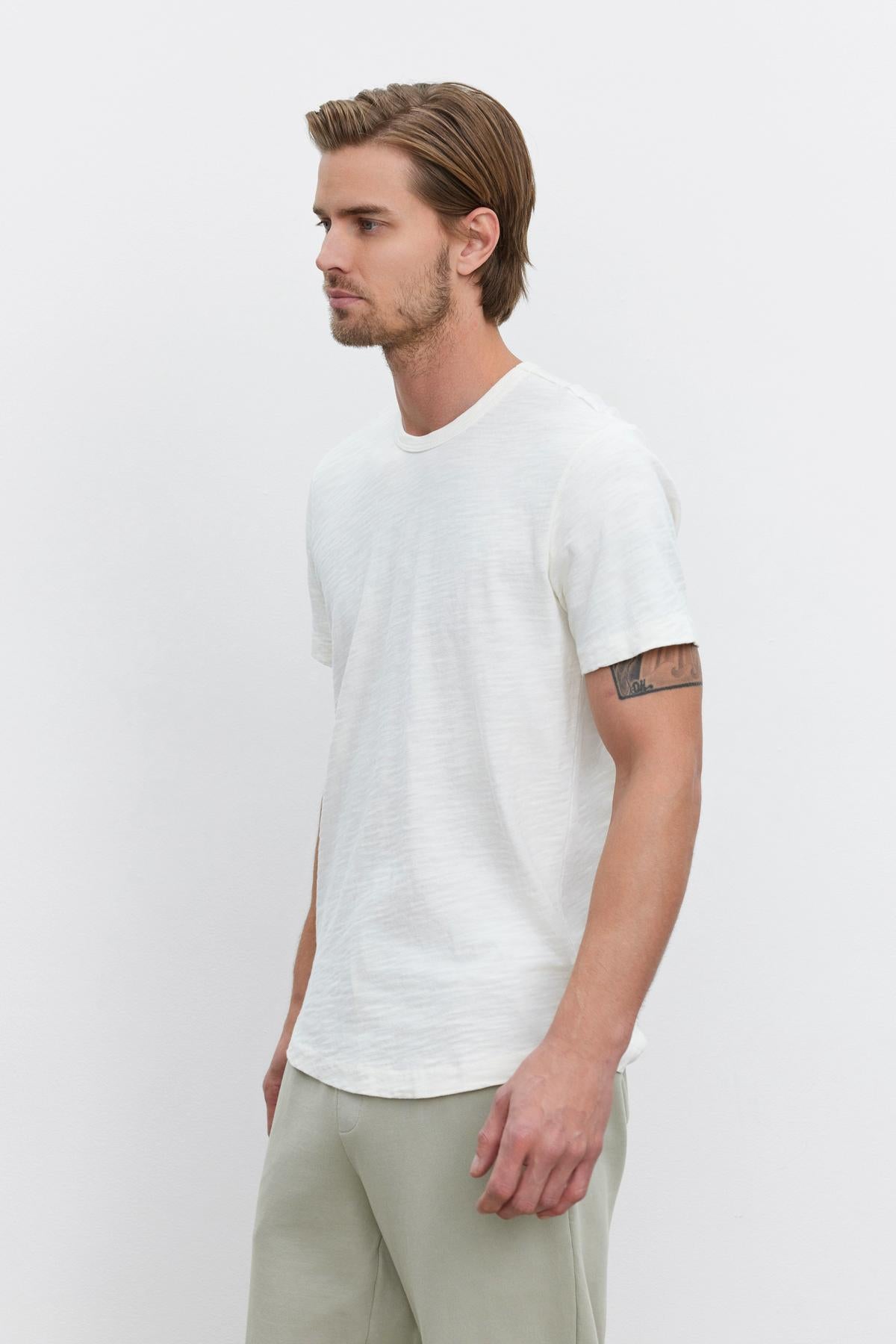   A man with short hair and a tattoo on his upper arm is standing against a plain white background, wearing a SPADER TEE by Velvet by Graham & Spencer and light-colored pants. 