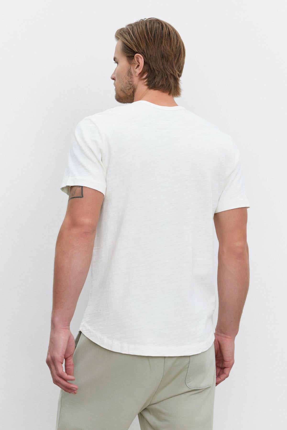   A man with short hair is shown from the back, wearing a white SPADER TEE by Velvet by Graham & Spencer and light grey pants. A tattoo is visible on his left upper arm. 