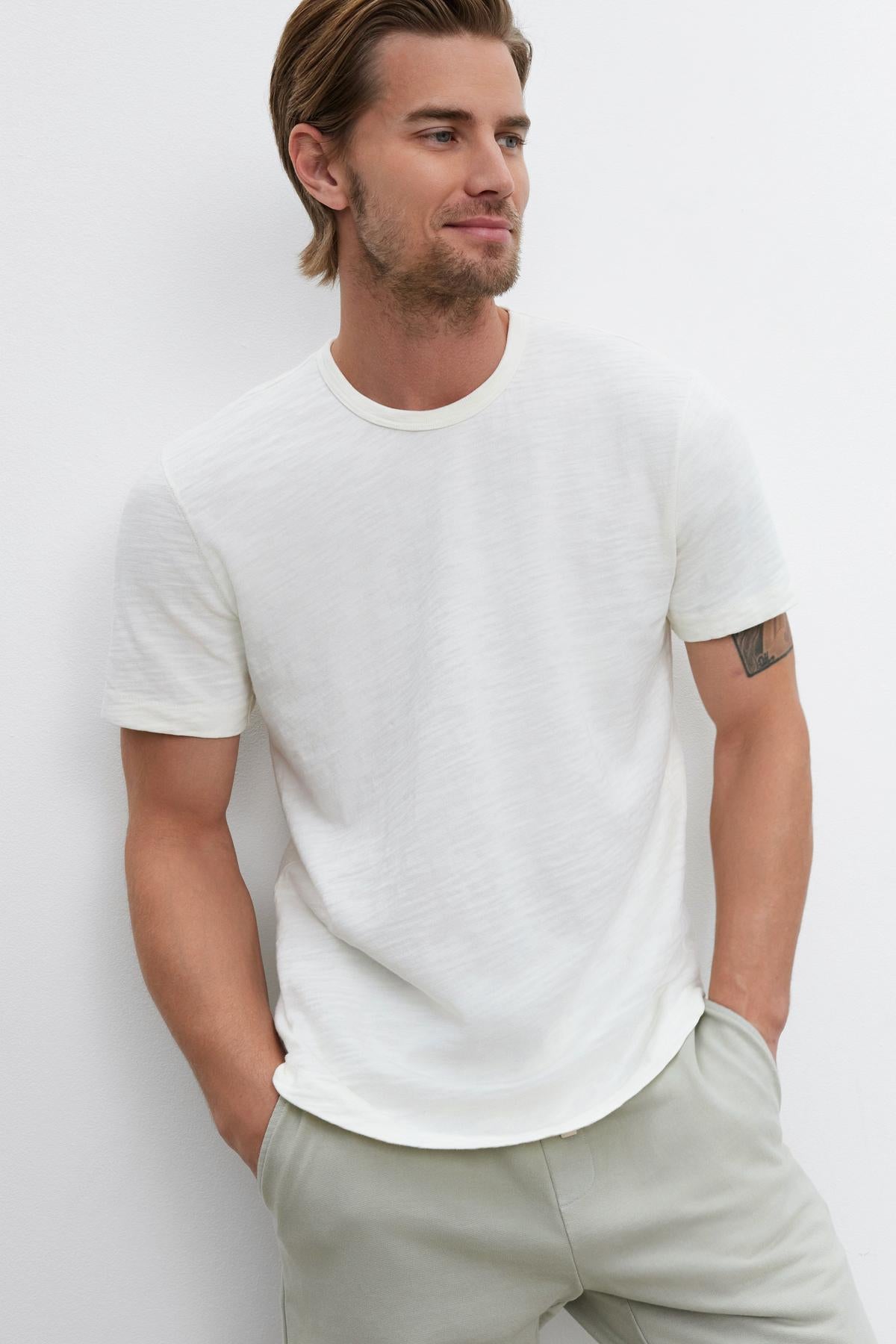   A man with light brown hair and a beard wearing a white SPADER TEE by Velvet by Graham & Spencer and light gray pants stands against a white background with his hands in his pockets. 