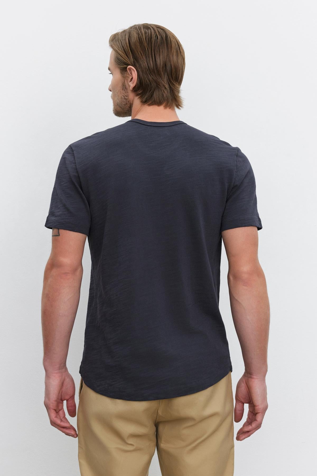   A man with light brown hair is wearing a dark gray SPADER TEE by Velvet by Graham & Spencer and beige pants, standing with his back to the camera against a plain white background. 