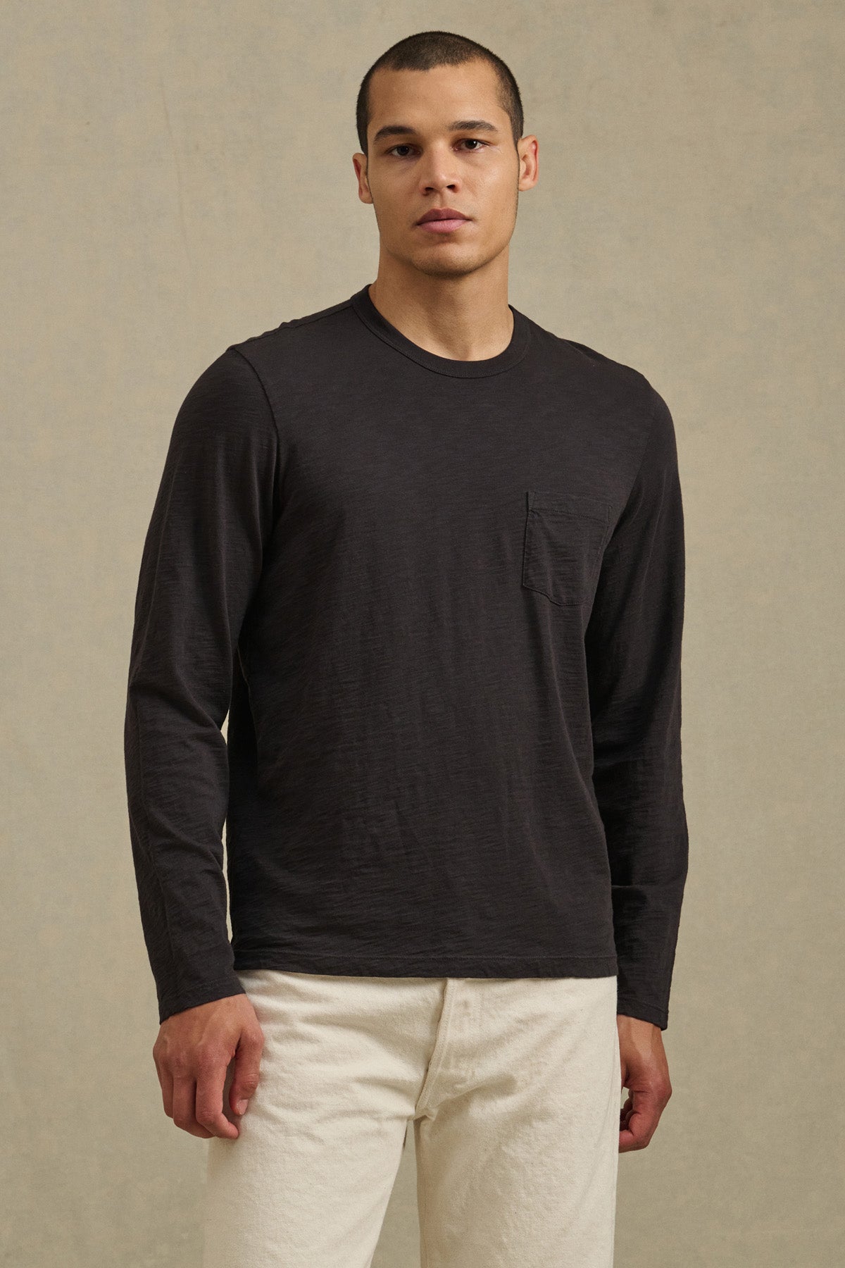 A man faces forward against a beige background, wearing the Velvet by Graham & Spencer DALTON TEE in black cotton slub with a chest pocket, paired with off-white pants.-40850714788033