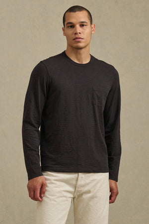 A man faces forward against a beige background, wearing the Velvet by Graham & Spencer DALTON TEE in black cotton slub with a chest pocket, paired with off-white pants.