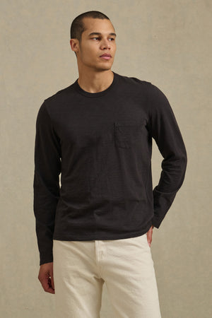 A man wears the DALTON TEE by Velvet by Graham & Spencer—a black cotton slub long-sleeve tee with a chest pocket—paired with off-white pants, standing against a neutral background for effortless everyday style.