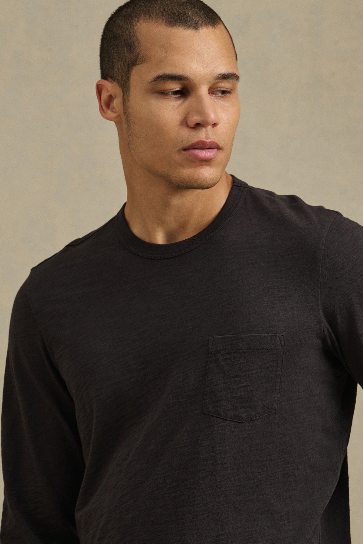 A man models the DALTON TEE by Velvet by Graham & Spencer—a black long-sleeve cotton slub tee with a chest pocket—against a neutral background, highlighting its effortless everyday style.