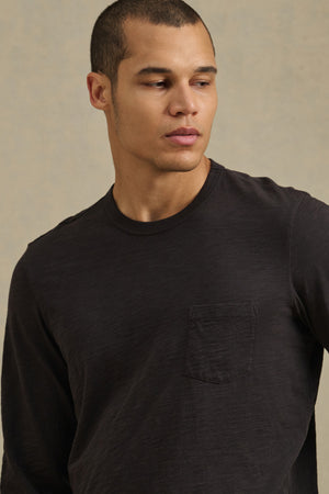 A man models the DALTON TEE by Velvet by Graham & Spencer—a black long-sleeve cotton slub tee with a chest pocket—against a neutral background, highlighting its effortless everyday style.