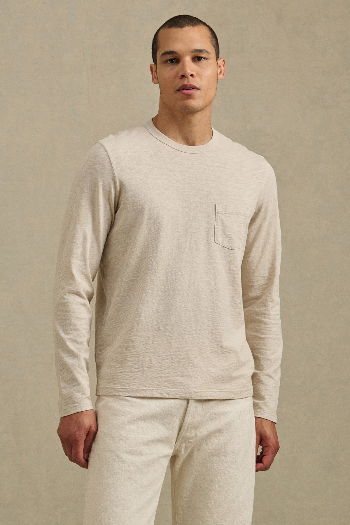 A man stands against a plain background wearing the DALTON TEE by Velvet by Graham & Spencer—a beige cotton slub long-sleeve shirt with a chest pocket and crew neckline—paired with matching beige pants.-41297077371073