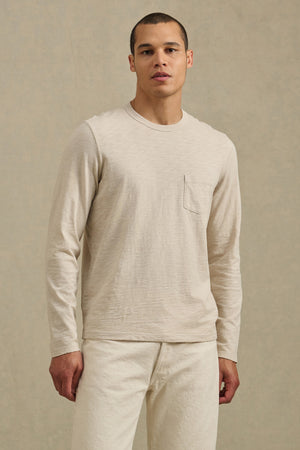 A man stands against a plain background wearing the DALTON TEE by Velvet by Graham & Spencer—a beige cotton slub long-sleeve shirt with a chest pocket and crew neckline—paired with matching beige pants.