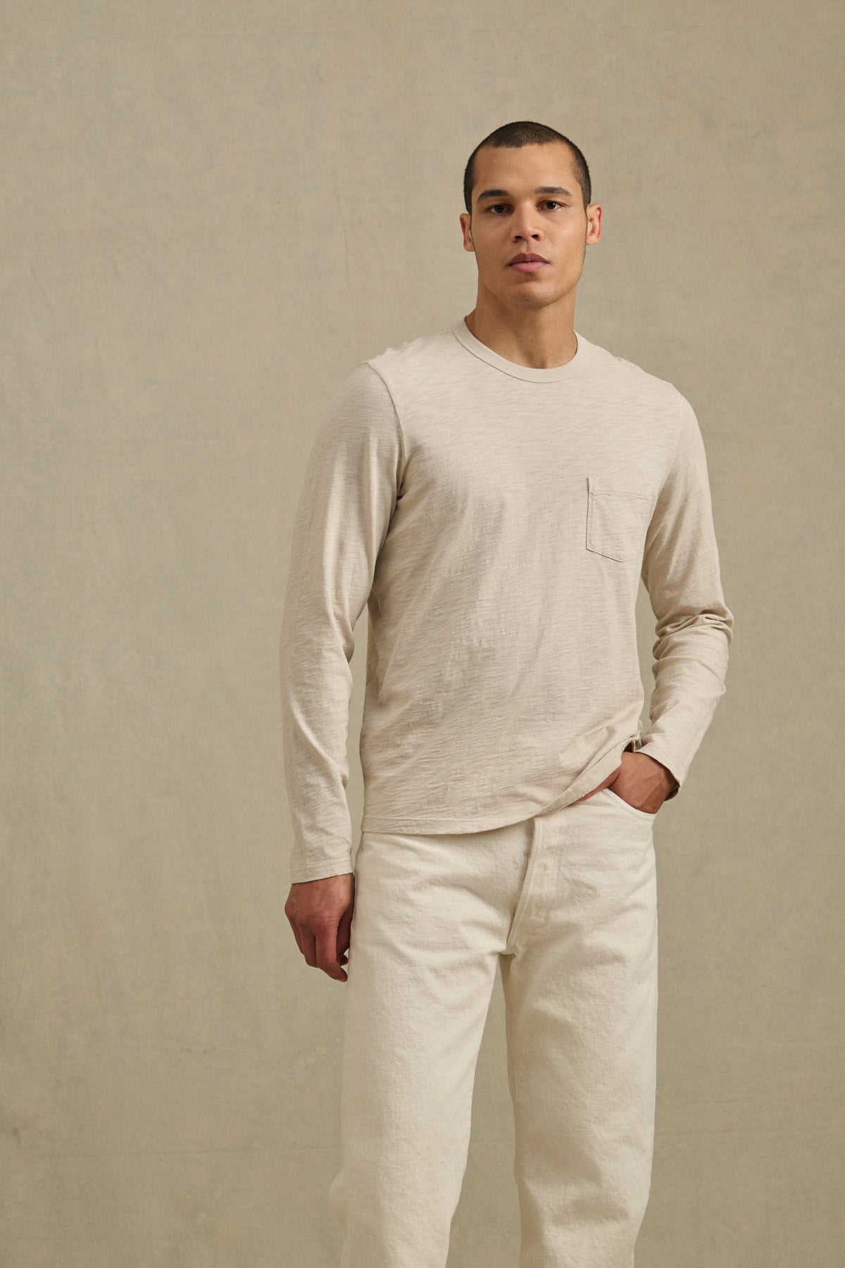 A man stands against a plain beige background, wearing the DALTON TEE by Velvet by Graham & Spencer—a cotton slub long-sleeve tee with a chest pocket and crew neckline—paired with cream pants.