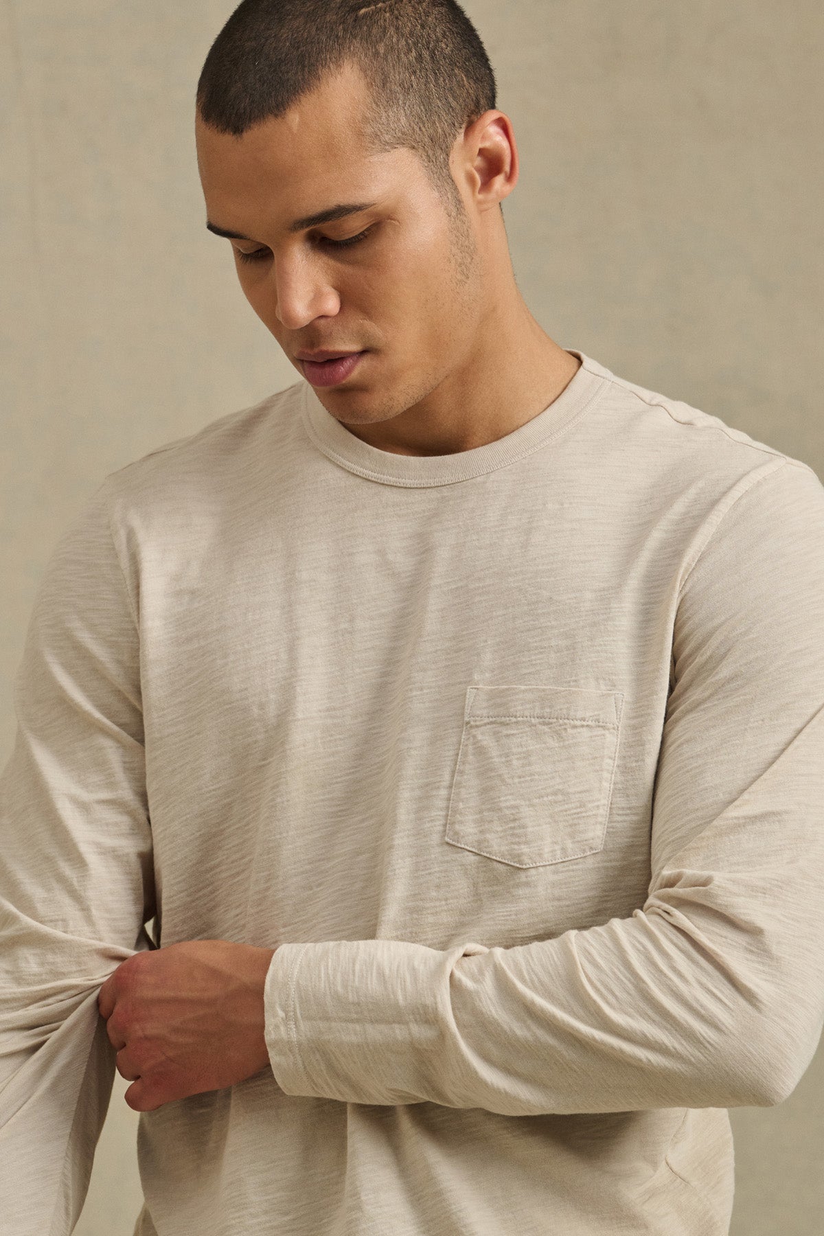 A man adjusts his sleeve while wearing the DALTON TEE by Velvet by Graham & Spencer, a light beige cotton slub long-sleeve shirt with a chest pocket and crew neckline.-41297077305537