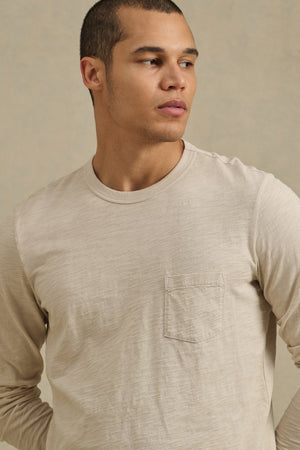 A person wears the DALTON TEE by Velvet by Graham & Spencer—a beige cotton slub long-sleeve shirt with a chest pocket and crew neck—standing against a neutral background, looking to the side.