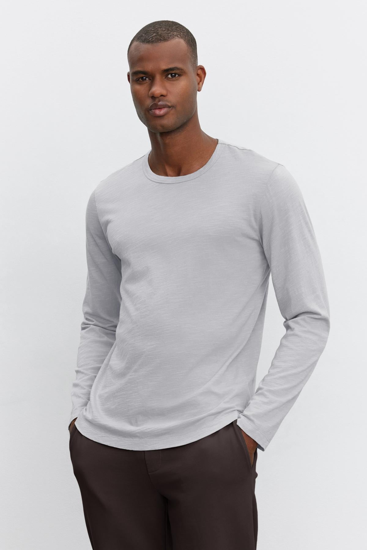   A man wearing the KAI TEE from Velvet by Graham & Spencer—a light gray long-sleeve shirt crafted from lightweight Peruvian cotton, featuring a classic crew neckline—and dark pants stands against a plain white background. 