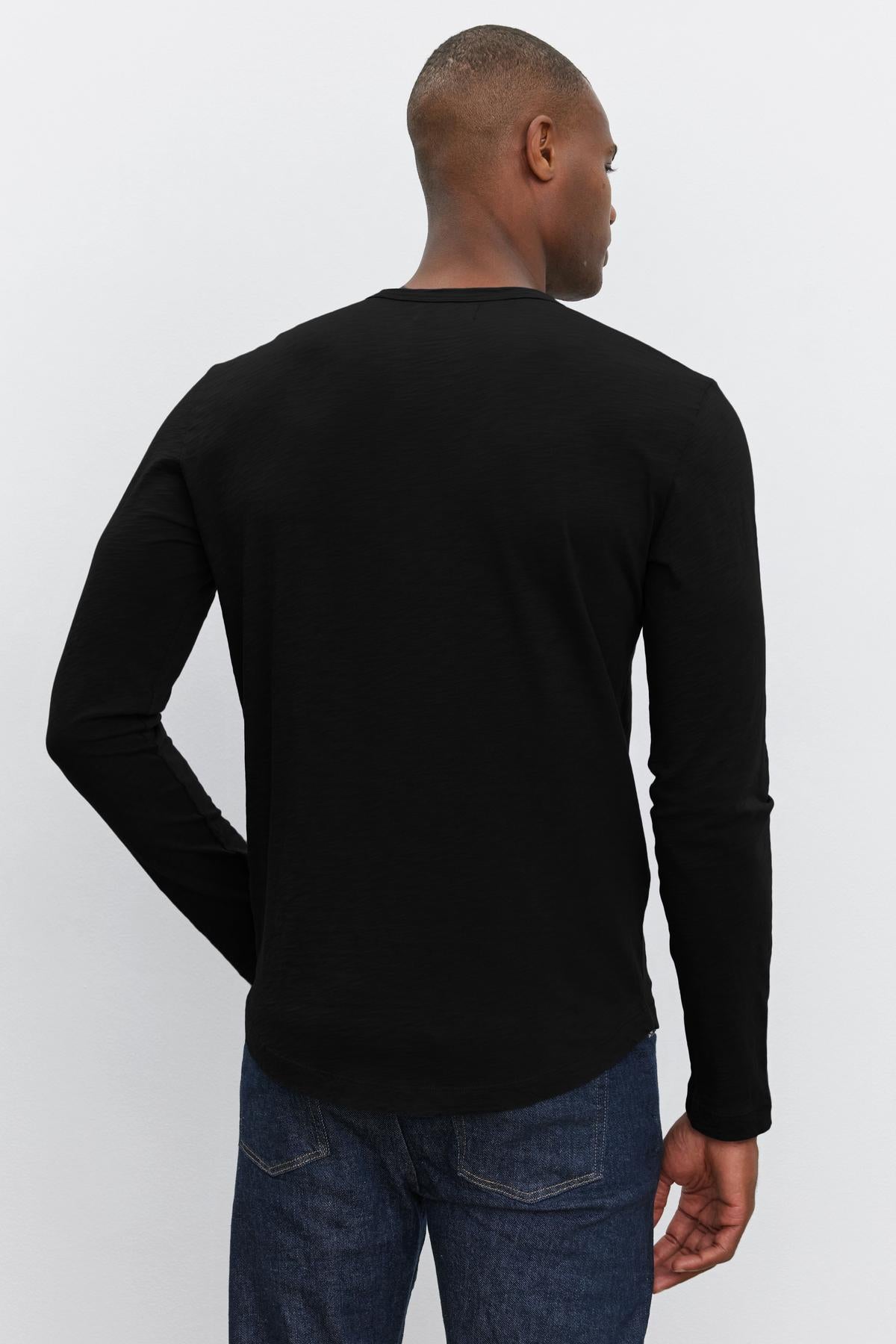   A person standing with their back to the camera, wearing a black long-sleeve KAI TEE from Velvet by Graham & Spencer featuring a classic crew neckline and dark jeans. 