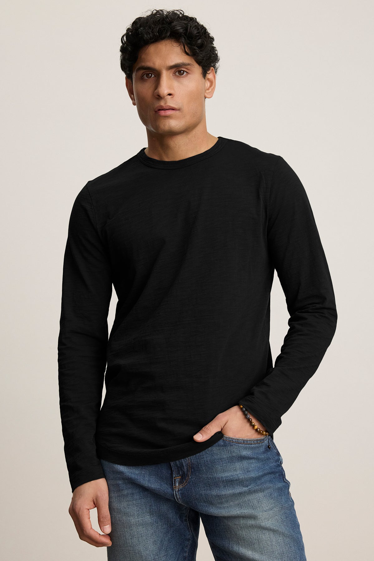 A curly-haired individual in a KAI TEE, a black long-sleeve shirt with a crew neckline by Velvet by Graham & Spencer, wears blue jeans and stands casually with one hand in their pocket.-38807449567425