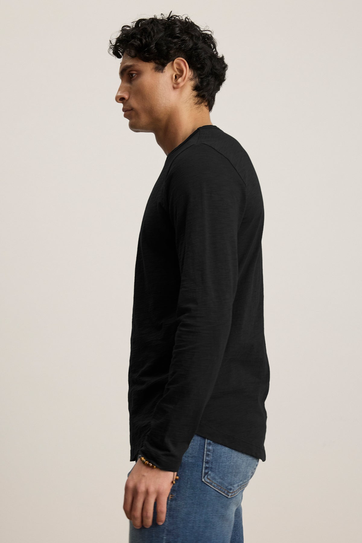   A person stands sideways against a light background wearing Velvet by Graham & Spencer's KAI TEE, a black long-sleeve shirt with a timeless design, paired with blue jeans. 